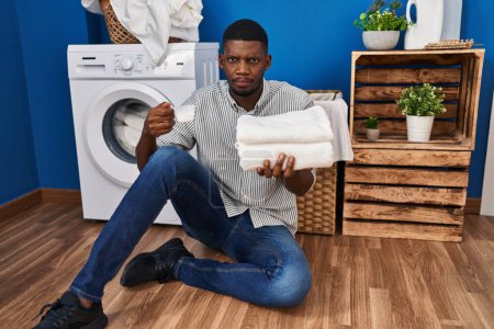 Téléchargez les photos : African american man holding clean laundry and laundry powder skeptic and nervous, frowning upset because of problem. negative person. - en image libre de droit
