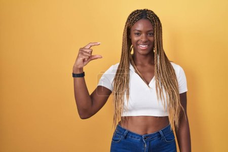 Téléchargez les photos : Femme afro-américaine avec les cheveux tressés debout sur fond jaune geste souriant et confiant avec la main faisant signe de petite taille avec les doigts regardant et la caméra. concept de mesure. - en image libre de droit