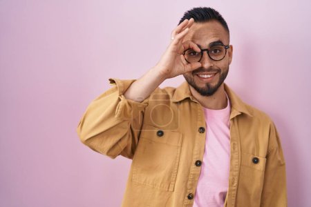 Foto de Young hispanic man standing over pink background doing ok gesture with hand smiling, eye looking through fingers with happy face. - Imagen libre de derechos