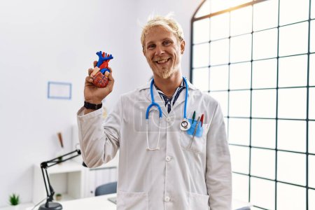Téléchargez les photos : Young blond man wearing doctor uniform holding heart at clinic looking positive and happy standing and smiling with a confident smile showing teeth - en image libre de droit