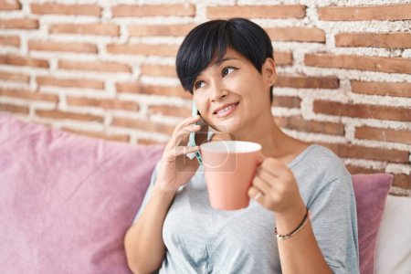 Téléchargez les photos : Middle age chinese woman talking on smartphone drinking coffee at bedroom - en image libre de droit