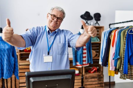 Téléchargez les photos : Senior man with grey hair working as manager at retail boutique approving doing positive gesture with hand, thumbs up smiling and happy for success. winner gesture. - en image libre de droit