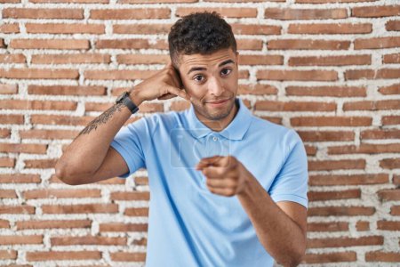 Téléchargez les photos : Jeune homme brésilien debout sur le mur de briques souriant faire parler au téléphone geste et vous pointant du doigt. Appelle-moi. - en image libre de droit