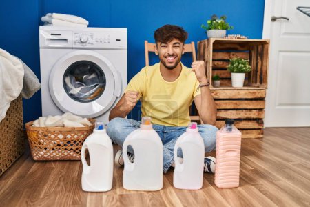 Téléchargez les photos : Arab man with beard doing laundry sitting on the floor with detergent bottle celebrating surprised and amazed for success with arms raised and eyes closed - en image libre de droit