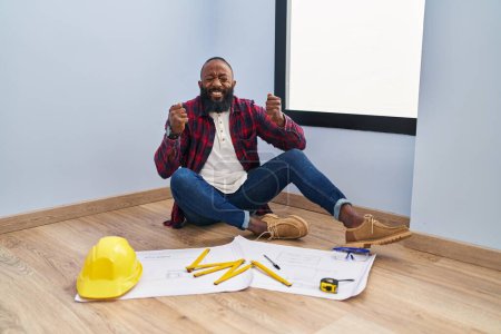Photo for African american man sitting on the floor at new home looking at blueprints excited for success with arms raised and eyes closed celebrating victory smiling. winner concept. - Royalty Free Image