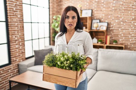 Foto de Mujer hispana joven sosteniendo plantas verdes en la sala de estar expresión despistada y confusa. concepto de duda. - Imagen libre de derechos