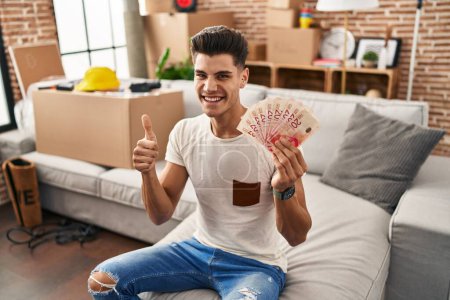 Foto de Young hispanic man moving to a new home holding shekels smiling happy and positive, thumb up doing excellent and approval sign - Imagen libre de derechos