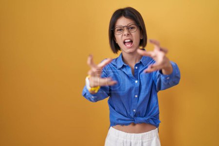 Photo for Young girl standing over yellow background shouting frustrated with rage, hands trying to strangle, yelling mad - Royalty Free Image