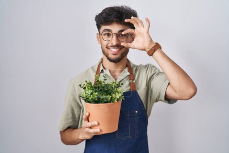 Téléchargez les photos : Arab man with beard holding green plant pot doing ok gesture with hand smiling, eye looking through fingers with happy face. - en image libre de droit