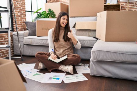 Foto de Young brunette woman moving to a new home doing finances smiling happy and positive, thumb up doing excellent and approval sign - Imagen libre de derechos
