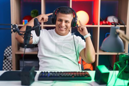 Téléchargez les photos : Jeune homme hispanique jouant à des jeux vidéo souriant pointant vers la tête avec les deux doigts des mains, grande idée ou pensée, bonne mémoire - en image libre de droit