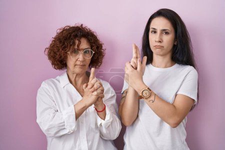 Téléchargez les photos : Hispanic mother and daughter wearing casual white t shirt over pink background holding symbolic gun with hand gesture, playing killing shooting weapons, angry face - en image libre de droit