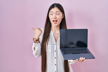 Foto de China joven mujer sosteniendo portátil que muestra la pantalla apuntando pulgar hacia el lado sonriendo feliz con la boca abierta - Imagen libre de derechos