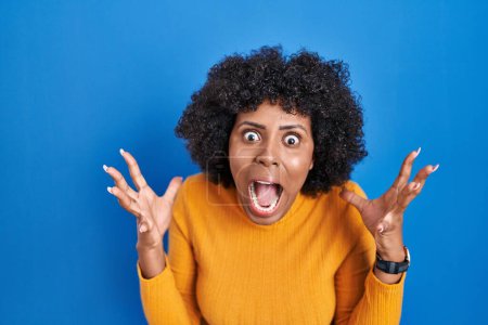 Photo for Black woman with curly hair standing over blue background crazy and mad shouting and yelling with aggressive expression and arms raised. frustration concept. - Royalty Free Image