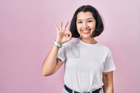 Foto de Mujer hispana joven vistiendo camiseta blanca casual sobre fondo rosa sonriendo positiva haciendo signo de ok con la mano y los dedos. expresión exitosa. - Imagen libre de derechos