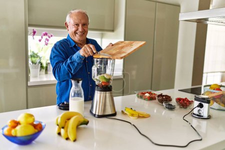 Téléchargez les photos : Senior homme souriant confiant mettre la banane dans le mélangeur à la cuisine - en image libre de droit