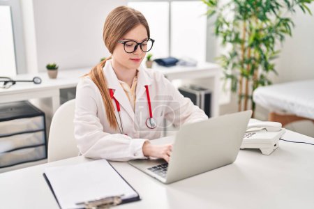 Téléchargez les photos : Young blonde woman wearing doctor uniform using laptop working at clinic - en image libre de droit