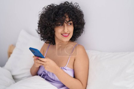 Téléchargez les photos : Jeune femme du Moyen-Orient utilisant smartphone assis sur le lit dans la chambre - en image libre de droit