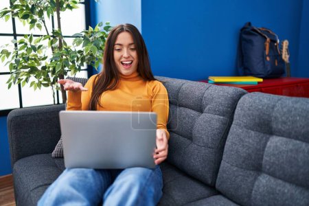 Photo for Young brunette woman working using computer laptop sitting on the sofa celebrating achievement with happy smile and winner expression with raised hand - Royalty Free Image