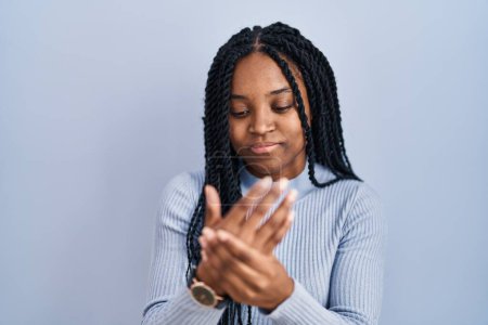 Foto de Mujer afroamericana de pie sobre fondo azul sufriendo dolor en manos y dedos, inflamación de la artritis - Imagen libre de derechos