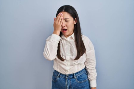 Foto de Young latin woman standing over blue background yawning tired covering half face, eye and mouth with hand. face hurts in pain. - Imagen libre de derechos
