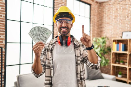 Foto de Joven hombre hispano con barba trabajando en la renovación del hogar sosteniendo dólares sonriendo con una idea o pregunta señalando con el dedo con cara feliz, número uno - Imagen libre de derechos