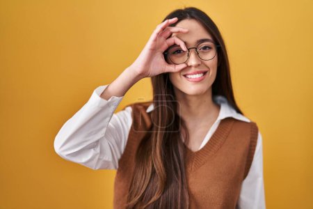 Photo for Young brunette woman standing over yellow background wearing glasses doing ok gesture with hand smiling, eye looking through fingers with happy face. - Royalty Free Image
