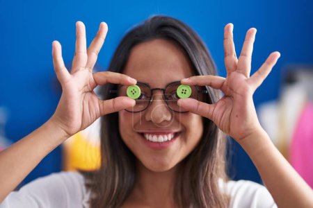 Téléchargez les photos : Young hispanic woman tailor smiling confident holding buttons over eyes at sewing studio - en image libre de droit
