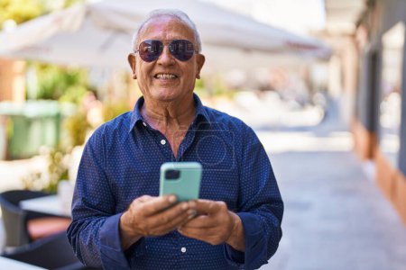 Téléchargez les photos : Homme âgé aux cheveux gris souriant confiant en utilisant un smartphone dans la rue - en image libre de droit