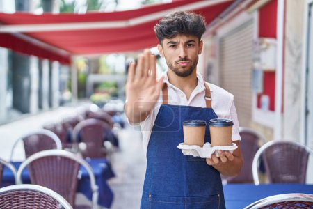 Téléchargez les photos : Arabe avec barbe portant tablier de serveur à la terrasse du restaurant avec la main ouverte faisant stop signe avec expression sérieuse et confiante, geste de défense - en image libre de droit