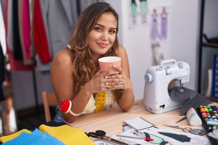 Téléchargez les photos : Jeune belle femme hispanique tailleur souriant boire du café en toute confiance au magasin de tailleur - en image libre de droit
