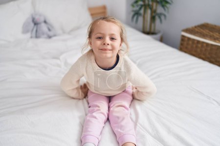 Téléchargez les photos : Adorable fille blonde souriante confiante assise sur le lit dans la chambre - en image libre de droit