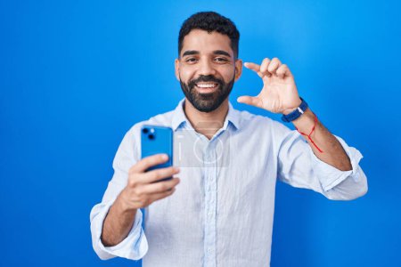 Téléchargez les photos : Hispanic man with beard using smartphone typing message smiling and confident gesturing with hand doing small size sign with fingers looking and the camera. measure concept. - en image libre de droit