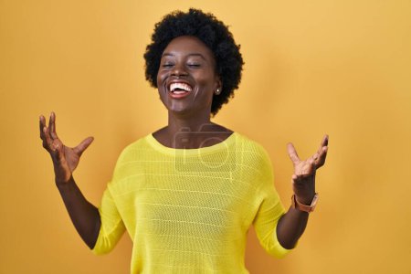 Foto de African young woman standing over yellow studio celebrating mad and crazy for success with arms raised and closed eyes screaming excited. winner concept - Imagen libre de derechos