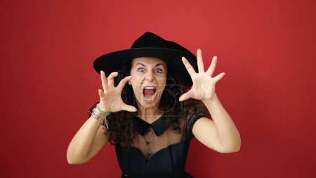 Foto de Mujer hispana de mediana edad con disfraz de bruja haciendo gesto de miedo con las manos sobre fondo rojo aislado - Imagen libre de derechos