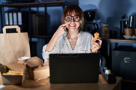 Foto de Young beautiful woman working using computer laptop and eating delivery food pointing with hand finger to face and nose, smiling cheerful. beauty concept - Imagen libre de derechos
