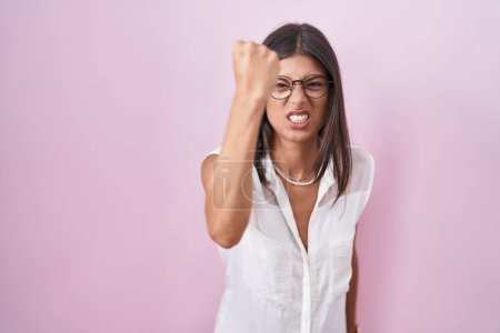 Foto de Mujer joven morena de pie sobre fondo rosa con gafas enojado y loco levantando el puño frustrado y furioso mientras gritaba con ira. rabia y concepto agresivo. - Imagen libre de derechos
