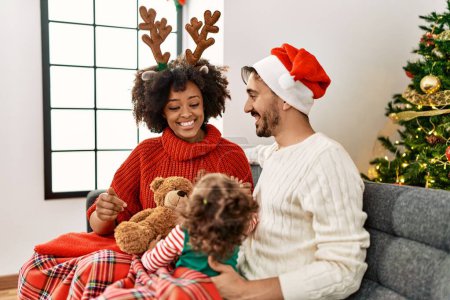 Foto de Couple and daughter playing with teddy bear sitting by christmas tree at home - Imagen libre de derechos