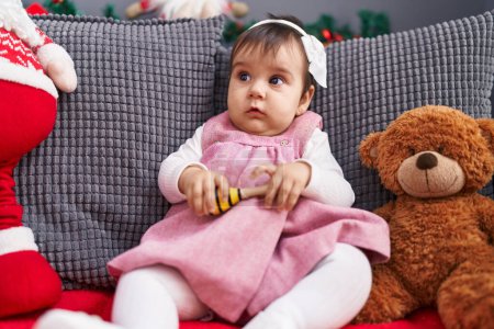Foto de Adorable bebé hispano jugando maraca sentado en el sofá por la decoración de Navidad en casa - Imagen libre de derechos