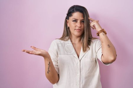 Foto de Blonde woman standing over pink background confused and annoyed with open palm showing copy space and pointing finger to forehead. think about it. - Imagen libre de derechos