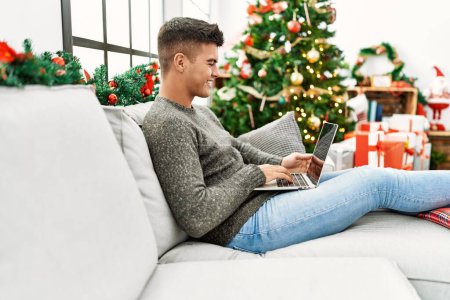 Photo for Young hispanic man using laptop sitting on sofa by christmas tree at home - Royalty Free Image