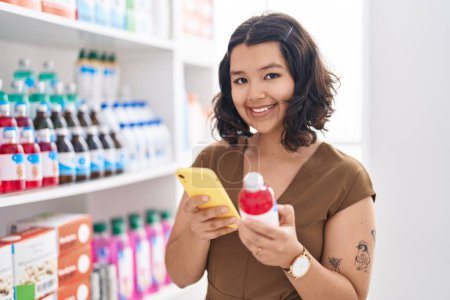 Photo for Young woman customer using smartphone holding medication bottle at pharmacy - Royalty Free Image