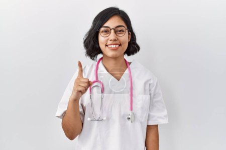 Foto de Young hispanic doctor woman wearing stethoscope over isolated background showing and pointing up with finger number one while smiling confident and happy. - Imagen libre de derechos