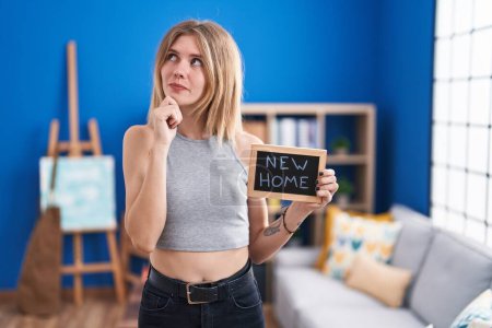 Photo for Blonde caucasian woman holding blackboard with new home text serious face thinking about question with hand on chin, thoughtful about confusing idea - Royalty Free Image