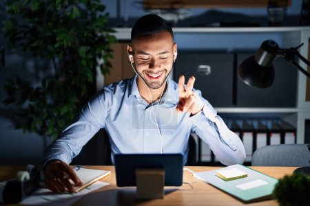 Photo for Young hispanic man working at the office at night smiling with happy face winking at the camera doing victory sign with fingers. number two. - Royalty Free Image