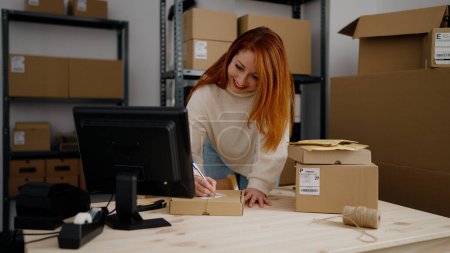 Photo for Young redhead woman ecommerce business worker writing on package at office - Royalty Free Image