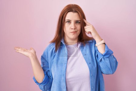 Téléchargez les photos : Young hispanic woman with red hair standing over pink background confused and annoyed with open palm showing copy space and pointing finger to forehead. think about it. - en image libre de droit