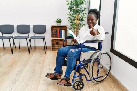 Téléchargez les photos : Jeune femme noire assise en fauteuil roulant dans la salle d'attente approuvant faire un geste positif avec la main, les pouces levés souriant et heureux pour le succès. geste gagnant. - en image libre de droit