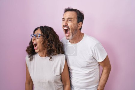 Foto de Middle age hispanic couple together over pink background angry and mad screaming frustrated and furious, shouting with anger. rage and aggressive concept. - Imagen libre de derechos