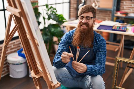 Foto de Young redhead man artist smiling confident holding paintbrushes at art studio - Imagen libre de derechos